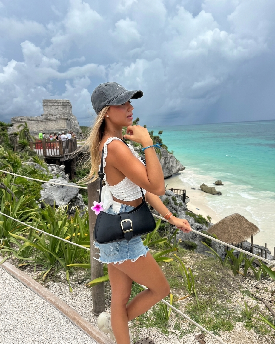 A stylish young woman wearing a Cyrycap baseball cap and vacation outfit, posing on a cliff overlooking the turquoise Caribbean sea at a beach resort.